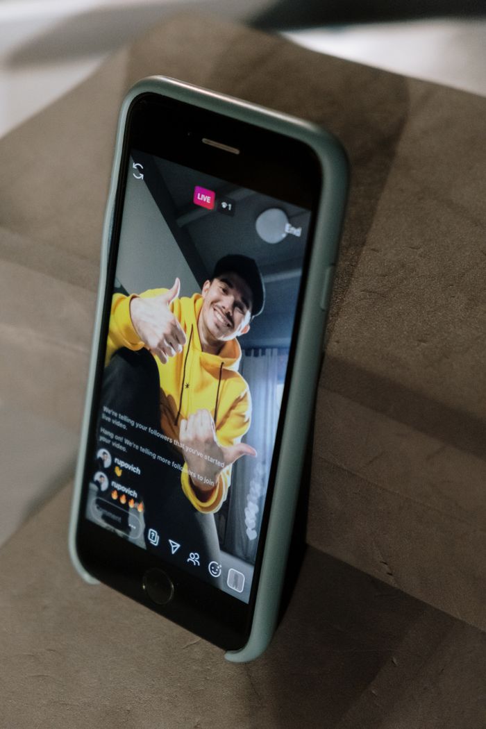 A man in a yellow hoodie taking part in a live stream on a smartphone screen, indoors.